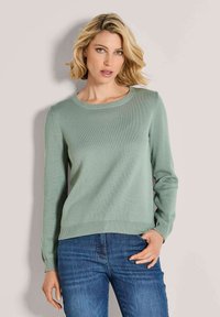 Frau mit kurzem blonden Haar, die einen hellgrünen Strickpullover und Jeans trägt, steht mit einer Hand in der Tasche vor einem einfarbigen Hintergrund.