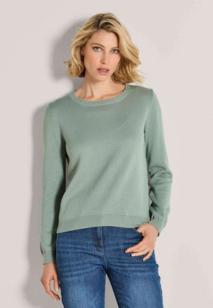 Frau mit kurzem blonden Haar, die einen hellgrünen Strickpullover und Jeans trägt, steht mit einer Hand in der Tasche vor einem einfarbigen Hintergrund.
