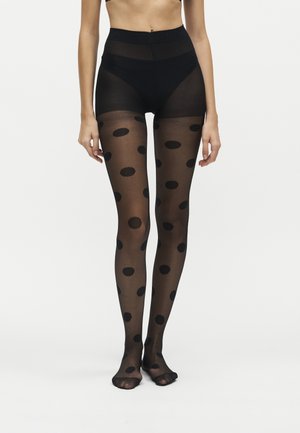 Femme portant des collants noirs transparents à gros pois noirs, debout devant un fond clair uni.