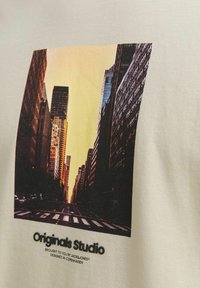 Lysebeige bomull T-skjorte med et trykk av en bygate ved solnedgang, med teksten "Originals Studio" i svart under bildet.
