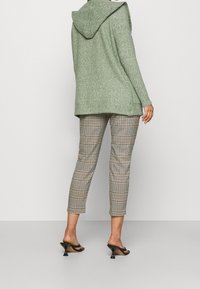 Zelený blejzr s kapucí z texturované látky, kombinovaný s kalhotami s hnědočernou kostkou. Nošen s černými sandály na podpatku.