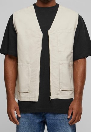 Uomo che indossa un gilet beige senza maniche con zip e tre tasche frontali sopra una maglietta nera a maniche corte e jeans blu su sfondo grigio.