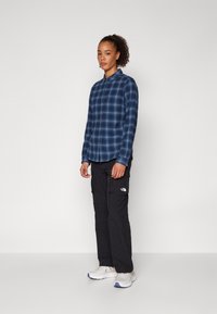 Camicia a maniche lunghe a quadri blu navy con chiusura a bottoni, abbinata a pantaloni cargo neri con tasche e scarpe da ginnastica grigie con accenti arancioni.