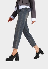 Scarpe nere con tacco a blocco in texture scamosciata e dettaglio a fiocco, abbinate a jeans in denim blu tagliati e sfilacciati e un elegante blazer grigio.