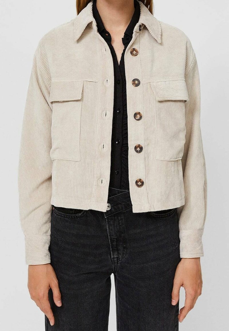 Femme portant une veste en velours côtelé beige à boutons par-dessus une chemise noire et un jean noir, sur un fond clair uni.