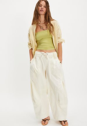 Νεαρή γυναίκα που φοράει ένα ανοιχτό κίτρινο ριγέ πουκάμισο πάνω από ένα πράσινο τοπ τύπου tube, φαρδιά κρεμ παντελόνια με κορδόνι, στρωμένα κολιέ και καφέ σανδάλια.