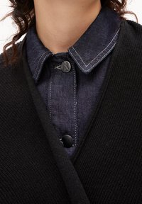 Svart stickad cardigan över en mörk denimskjorta med knapp-up krage. Har kontrasterande sömmar på kragen och knapparna.