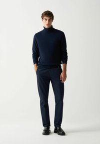 Jūras zilas krāsas turtleneck džemperis kopā ar tumši jūras zilas krāsas biksēm, to raksturo gluda tekstūra. Melni apavi noslēdz apģērbu. Vienkāršs un moderns dizains.