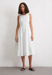 Robe blanche en lin longueur genoux avec un col rond et sans manches ; présente une silhouette fluide et une texture subtile. Sandales plates noires.