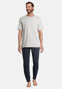 Mies, który się uśmiecha, z brodą, ubrany w jasnoszary t-shirt z krótkim rękawem i ciemnogranatowe spodnie dresowe, stoi boso na jednolitym tle.