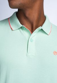 Mężczyzna w jasnozielonej koszulce polo z koralowymi wykończeniami kołnierzyka i małym koralowym logo na piersi, pokazany od szyi do połowy torsu.