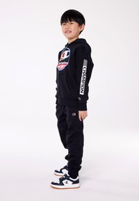 Completo felpa con cappuccio e pantaloni neri caratterizzato da un logo prominente, polsini a costine e un design sportivo. Completato da sneakers nere e bianche con suole in gomma.