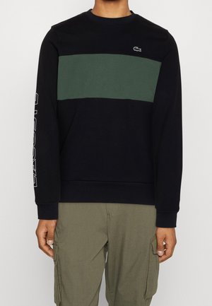Mand iført en sort langærmet sweatshirt med en bred, mørkegrøn horisontal stribe og khakifarvede cargobukser, stående mod en hvid baggrund.