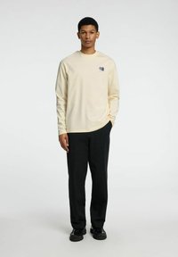 Långärmad ljusbeige bomullsskorta med logodetalj, kombinerad med svarta byxor och svarta skor, med en avslappnad passform och enkel design.