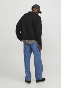 Schwarze übergroße Jacke mit Kragen, getragen über einem grau karierten Hemd und blauen lässigen Jeans. Eine schwarze Mütze und dunkle Schuhe vervollständigen das Outfit.