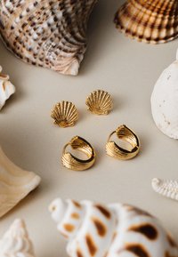 Gullformede øreringer i gull og matchende studs har teksturert detaljer. Omringet av forskjellige skjell på en lysecrème bakgrunn.