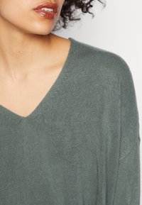 Grøn striktrøje med V-hals og blødt materiale, der har løstsiddende ærmer og et afslappet design. Detalje af halsudskæringen.