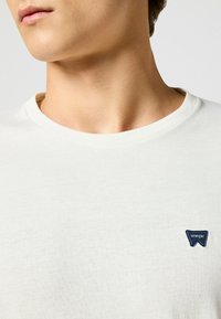 Biała bawełniana koszulka z okrągłym dekoltem, z małą niebieską naszywką z logo "Wrangler" na lewej piersi, gładka w dotyku.