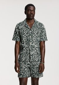 Un set composto da camicia a maniche corte con bottoni e pantaloncini realizzato in tessuto decorato in verde scuro e bianco. Presenta un motivo astratto di foglie su tutta la superficie.