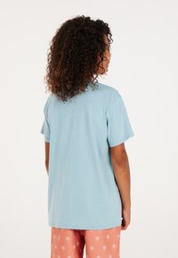 lichtblauwe t-shirt met korte mouwen en een ontspannen pasvorm. Gladde textuur, ronde hals, gedragen met roze pyjamabroek met witte palmbomenprint.