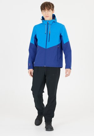 Blauwe en marineblauwe waterdichte jas met een chevronontwerp, verstelbare capuchon en ritszakken. Draagt met zwarte broek en laarzen.