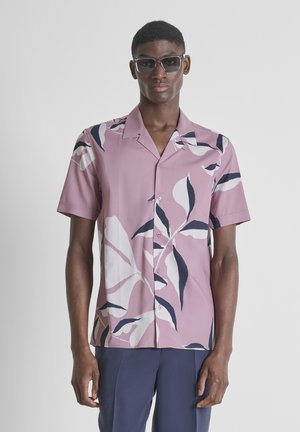 Muškarac u ljubičastoj košulji s kratkim rukavima i bijelim te mornarsko plavim uzorkom listova, mornarsko plavim hlačama i tamnim kvadratnim sunčanim naočalama, stoji na sivom zidu.