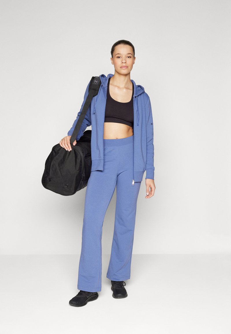 Felpa blu con zip frontale, pantaloni tuta blu coordinati, reggiseno sportivo nero, borsa a tracolla nera e scarpe nere, su uno sfondo semplice.