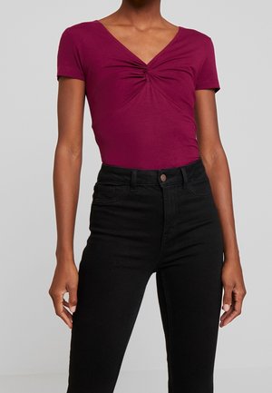 Femme portant un top froncé rose profond à manches courtes avec un col en V, associé à un pantalon noir taille haute sur un fond uni.