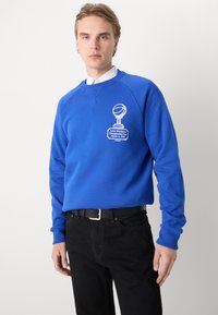 Blauwe sweatshirt van katoen met een ronde halslijn en lange mouwen. Kenmerkt zich door een witte bedrukt grafiek en tekst op de voorkant. Gekleed met zwarte broek.