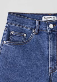 Ceinture de jeans en denim bleu avec bouton argenté et rivets, poches avant, passants pour ceinture, et étiquette de taille Pull&Bear à l'intérieur de la ceinture.