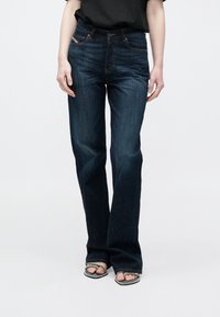 Σκούρα μπλε τζιν με φαρδιά καμπάνα από τζιν υλικό, με το κλασικό σχέδιο πέντε τσεπών, εμφανείς ραφές και ελαφριά σκιάσεις στο εμπρός μέρος.