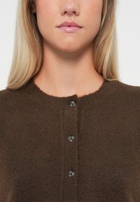 Καφέ κοντό πουλόβερ με στρογγυλή λαιμόκοψη, ribbed άκρες και πέντε σκούρα κουμπιά μπροστά. Μαλακή υφή με άνετη εμφάνιση.