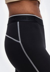 Κοντινό πλάνο ενός ατόμου που φοράει μαύρο κολάν Under Armour με γκρι ραφές αντιθέσεως και ορατή ζώνη μέσης με το λογότυπο της μάρκας.