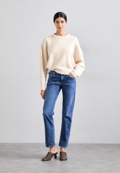 Suéter de lana suave color beige claro, jeans rectos azules y mules marrones texturizados con un tacón redondeado. Diseño simple y relajado con materiales suaves.
