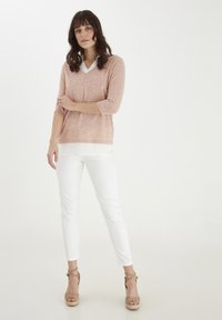 Lyserød tekstureret sweater med V-hals, lagd oven på en hvid skjorte, parret med hvide tætsiddende bukser og brune snorede kile-sandaler.
