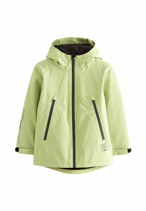 Osoba ubrana w jasnozieloną kurtkę zapinaną na zamek z czarnymi kieszeniami na zamek, na czarnej koszuli i ciemnych spodniach cargo, stojąca na jednolitym tle.