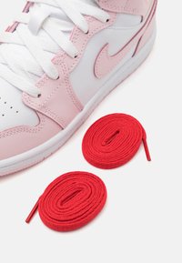 Baltas sneakers ar rozā akcentiem, ar teksturētu ādas augšdaļu, baltām šņorēm un divām sarkanām apaļām šņorēm, kas novietotas zemāk.