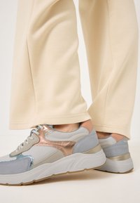 Sneaker grigi e argentati con texture in rete e suede, caratterizzati da dettagli in oro rosa e una suola bianca, abbinate a pantaloni color crema.