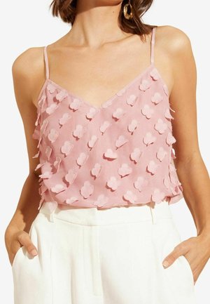 Kvinde iført en lyserød ærmeløs V-hals top med teksturerede blomstapplikationer og hvide bukser med høj talje, hænderne i lommerne.