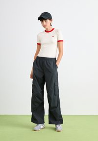 T-shirt crème ajustée avec des bordures rouges, associée à un pantalon cargo noir et des baskets grises, sur fond clair avec un sol vert.