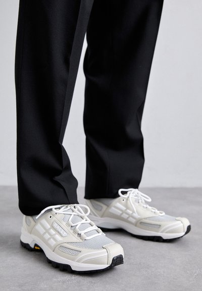 Adidași albi din plasă și material sintetic, cu accente gri, având un vârf texturat și șireturi albe. Pantaloni negri parțial vizibili.