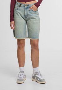 Persona vistiendo un pantalón corto de mezclilla azul claro con dobladillo sin terminar, calcetines blancos, zapatillas grises de Adidas y una camiseta corta de manga larga color burdeos, con una mano en el bolsillo.