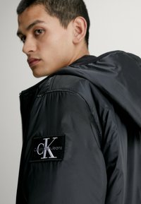 Giacca a vento nera con cappuccio, dotata di un patch del logo Calvin Klein Jeans sul lato sinistro, realizzata in tessuto lucido e liscio.