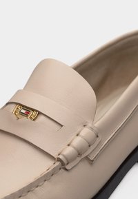 Μπεζ δερμάτινο μοκασίνι με μπροστινό λουράκι που φέρει ένα μικρό χρυσό έμβλημα και λεπτομερή ραφή κατά μήκος του άνω άκρου.