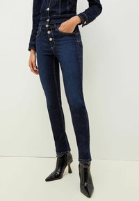 Mörkblå denim skinny jeans med metallknappstängning, femficksdesign och rena, avsmalnande ben, i kombination med svarta ankelboots.