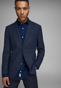Blazer blu navy in tessuto strutturato, con due bottoni e tasche a patta, indossato sopra una camicia blu a fantasia con bottoni. Jeans scuri completano l'outfit.