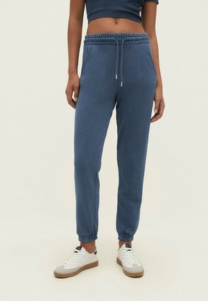 Kvinde iført blå joggebukser med snøre og elastiske manchetter og hvide sneakers, stående mod en ensfarvet baggrund.