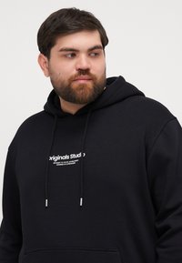 Svart hettegenser med snøring, med et hvitt logo som sier "Originals Studio" på brystet. Mykt stoff med en avslappet passform.