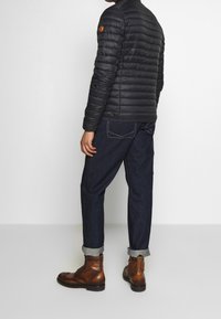 Giacca a vento nera con design trapuntato, abbinata a pantaloni in denim e stivali marroni con lacci. Presenta polsini arrotolati e un sottile dettaglio di branding.