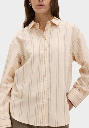 Mujer con camisa beige holgada de botones y finas rayas verticales, posando con una mano en la cadera sobre un fondo liso.
